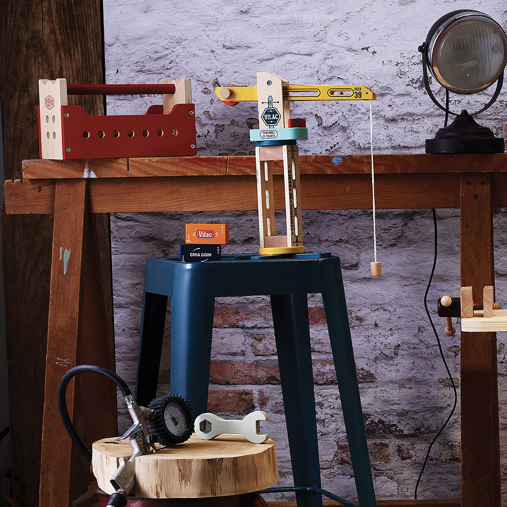 Ein Container-Kran aus Holz des Produkts 'Vilacity' von Vilac, gezeigt auf einem rauen, industriellen Hintergrund. Das Set enthält diverse farbenfrohe Bauteile, wie einen roten Hebekran, einen blauen Containerständer und kleine Accessoires, die die Vorstellungskraft und Feinmotorik von Kindern fördern.