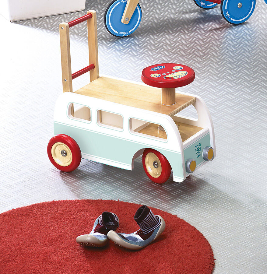 Holzspielzeug in Form eines Minibusses mit roten und weißen Details auf einem roten Teppich, daneben ein Paar Kinderhausschuhe.