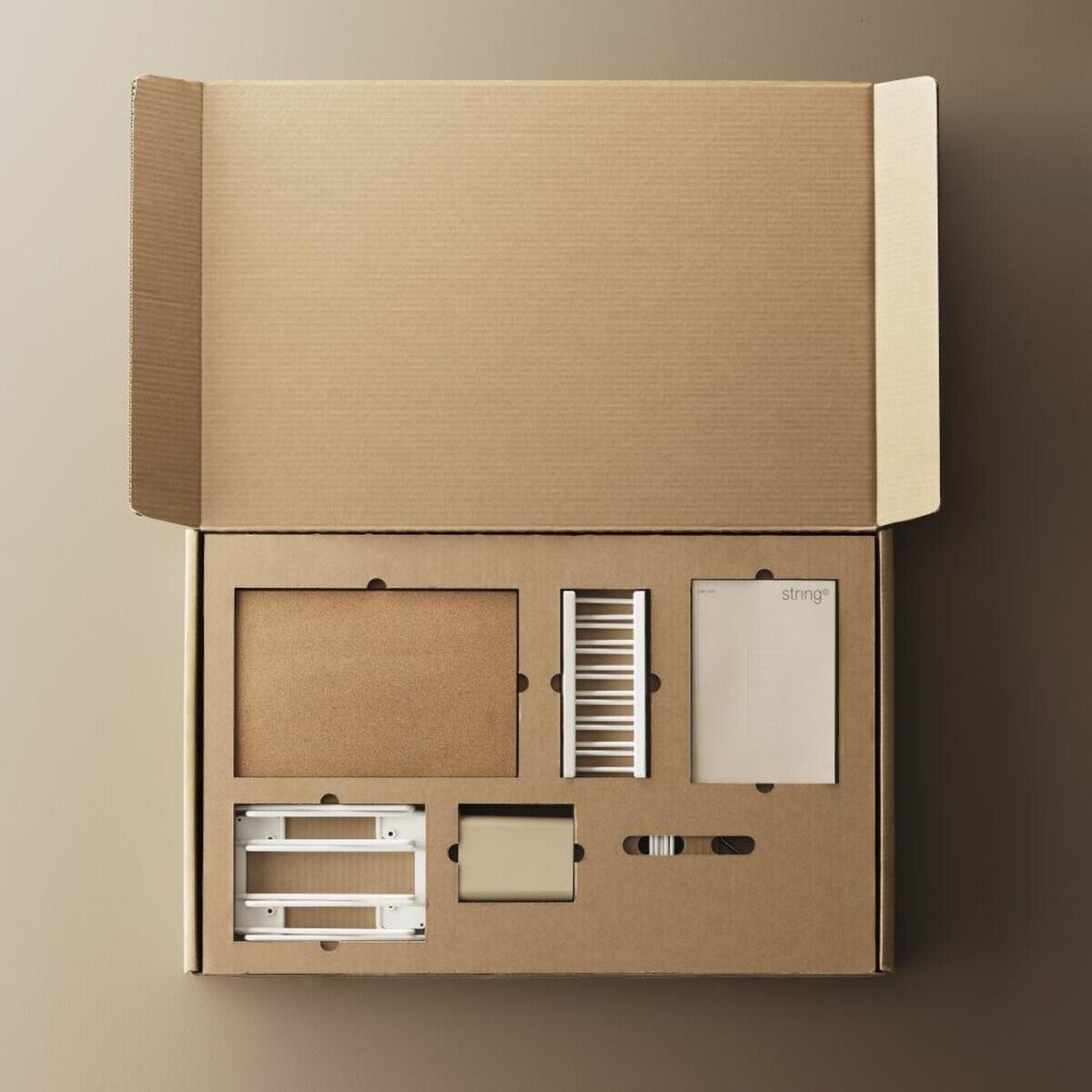 Ein offener Karton mit den Einzelteilen des String Küchenregals, einschließlich Regalböden, Halterungen und Montagematerialien. Der Karton und die Teile sind in neutralen Brauntönen gehalten.