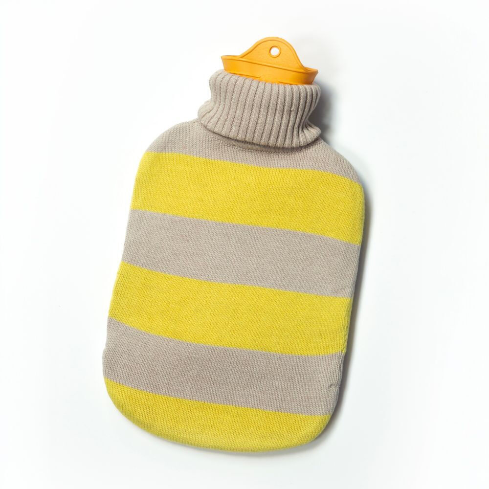 Freigestellte SUITE702 Wärmflasche »gestreift« vor weißem Hintergrund. Die ovale Wärmflasche trägt einen weich gestrickten Bezug mit breiten gelben und beigegrauen Querstreifen sowie geripptem, hohem Kragen. Oben ragt ein orangefarbener Latexverschluss mit kleiner Öse heraus.