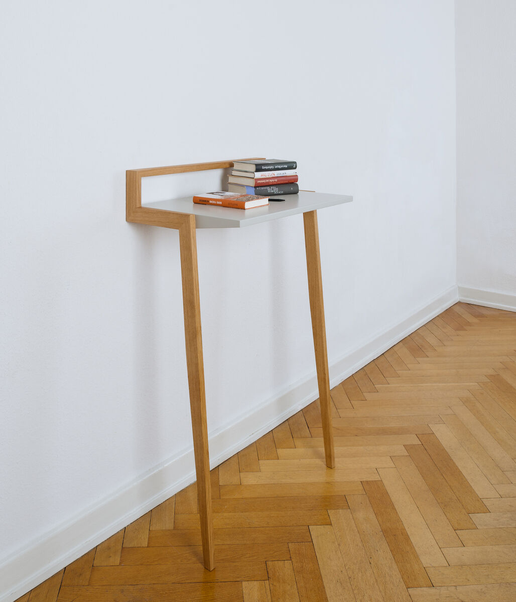 Ein modernes und minimalistisch gestaltetes Stehpult namens »buckowski« aus Holz mit weißer Tischplatte und schmalem Design, das Bücher und Dekorationen trägt. Es steht in einem Raum mit hellem Holzboden und weiß gestrichenen Wänden.