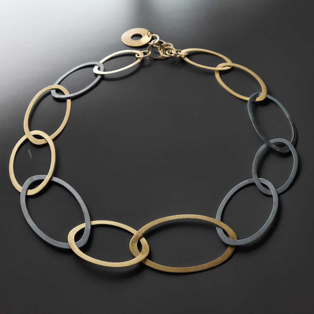 Zu sehen ist das Collier »Silberringe« von Maria Rzewuska Designerschmuck. Die Halskette besteht aus ineinandergreifenden, teils oxidierten und teils Feingold plattierten 925er Silberelementen in ovaler Form, angeordnet auf dunklem Hintergrund.