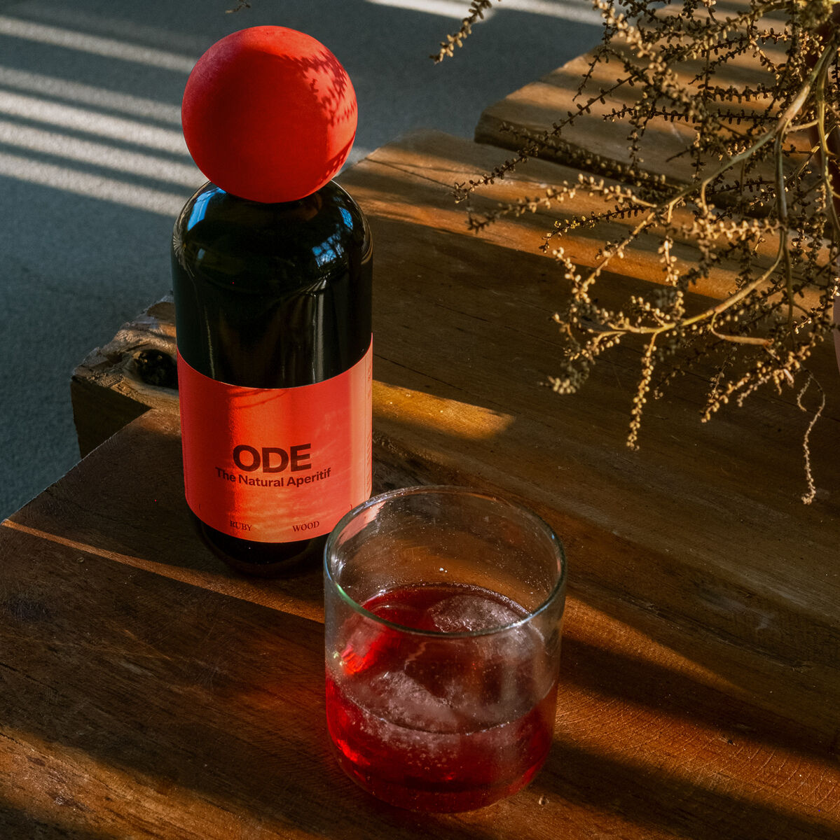 Eine Flasche Aperitif 'Ruby Wood' steht auf einem Holztisch neben einem Glas mit rotem Getränk und Eiswürfeln. Im Hintergrund ist eine Pflanze zu sehen, beleuchtet von Sonnenstrahlen.
