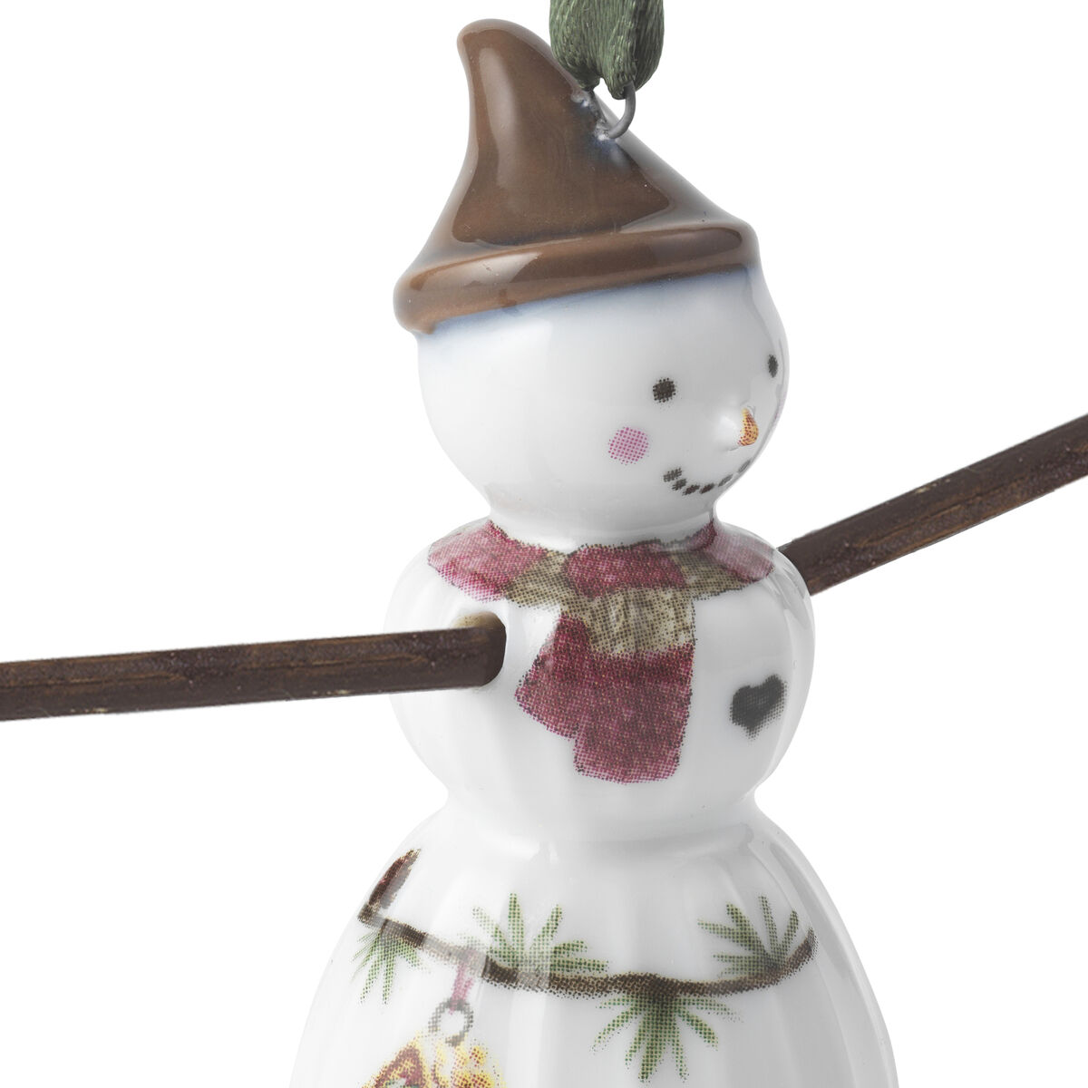 Porzellananhänger in Form eines Schneemädchens mit rot-braunem Schal, brauner Mütze und aufgemaltem Herz auf der Brust. Das Schneemädchen hält einen braunen Ast, an dem es befestigt ist. Auf dem weißen Kleid sind grüne Zweige und ein kleines gelbes Vogelhäuschen abgebildet. Der Anhänger eignet sich als weihnachtliche Dekoration oder Christbaumschmuck.