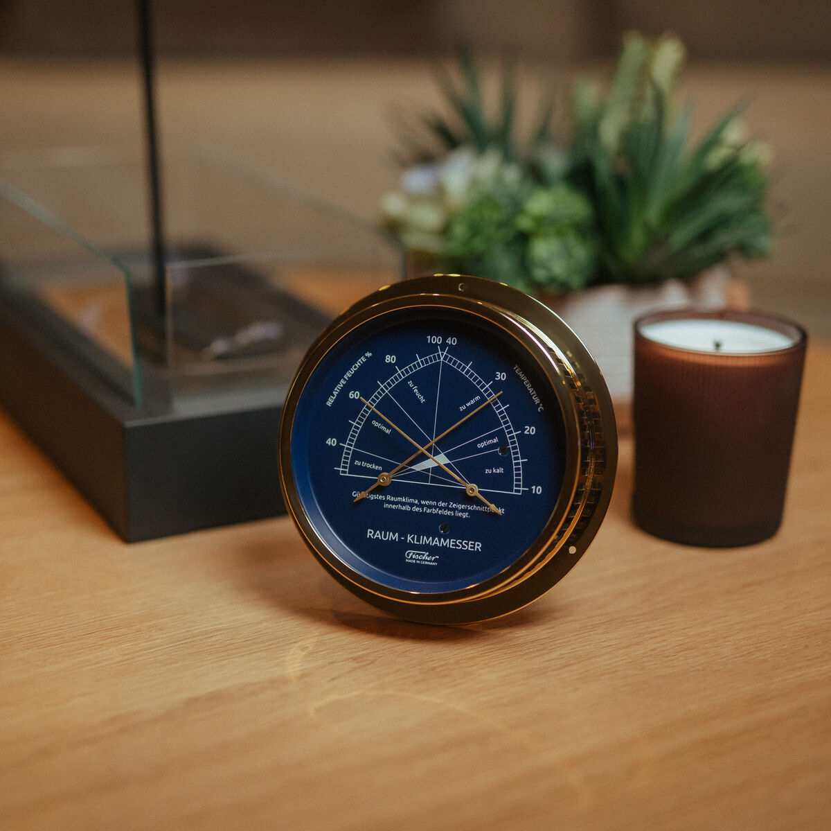 Das Bild zeigt ein ZEIT Sonderedition Raumklimamesser mit poliertem Messinggehäuse und blauem Zifferblatt. Das Gerät steht auf einem Holztisch, im Hintergrund sind eine braune Kerze, ein modernes Deko-Objekt und eine Pflanze zu sehen. Die Szene wirkt wohnlich und stilvoll, mit Fokus auf dem eleganten Messinstrument.