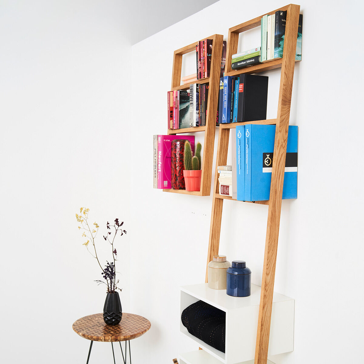 Ein modernes Standregal aus Holz, dekoriert mit Büchern und Pflanzen, steht in einem hellen Raum neben einem kleinen Tisch mit einer Vase und Blumen.