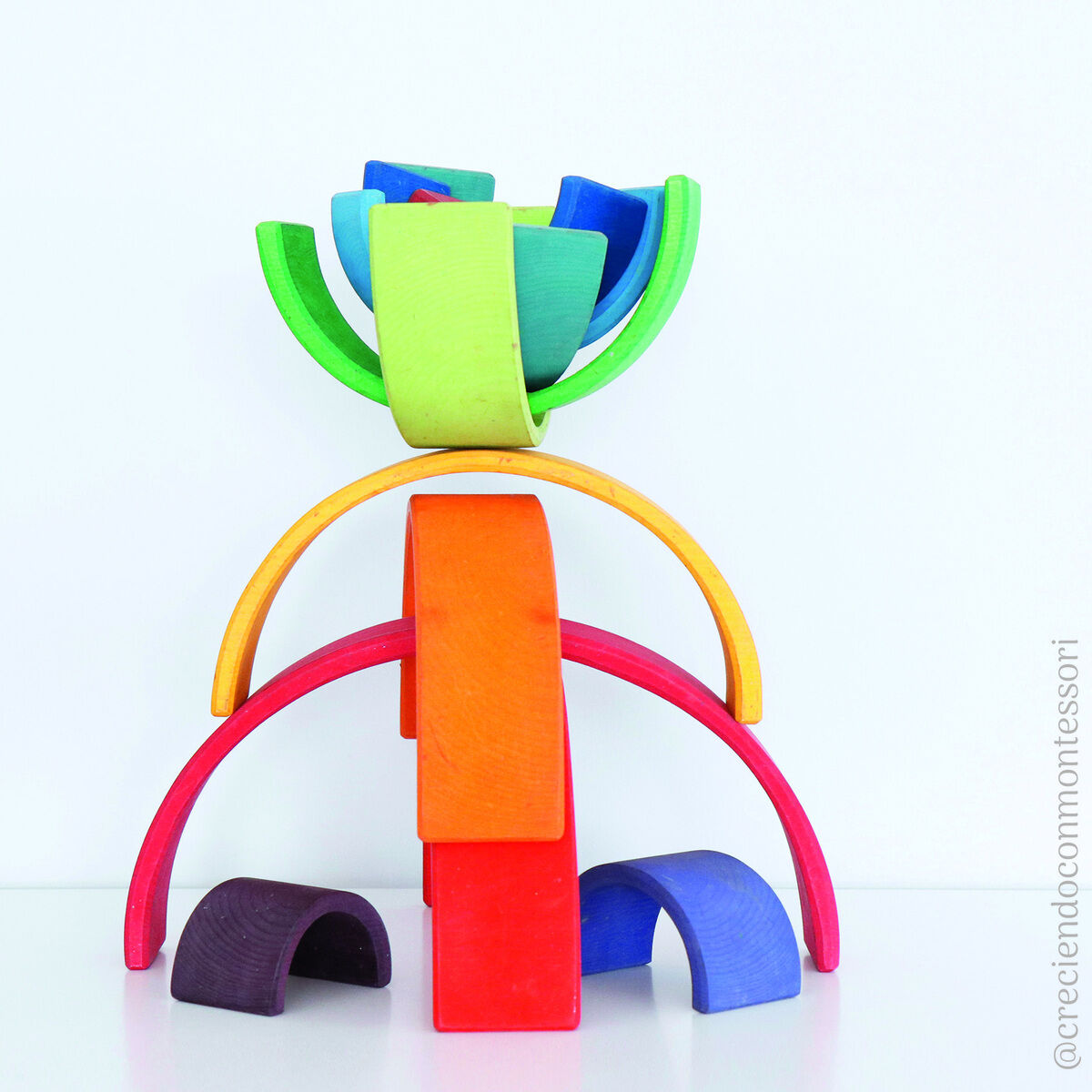 Ein Regenbogen-Holzspielzeug bestehend aus gebogenen, bunt bemalten Holzstücken, die zu einer kreativen Stapelstruktur angeordnet sind. Die Teile sind in Rottönen, Orangetönen, Gelbtönen, Grüntönen, Blautönen und Violetttönen bemalt.