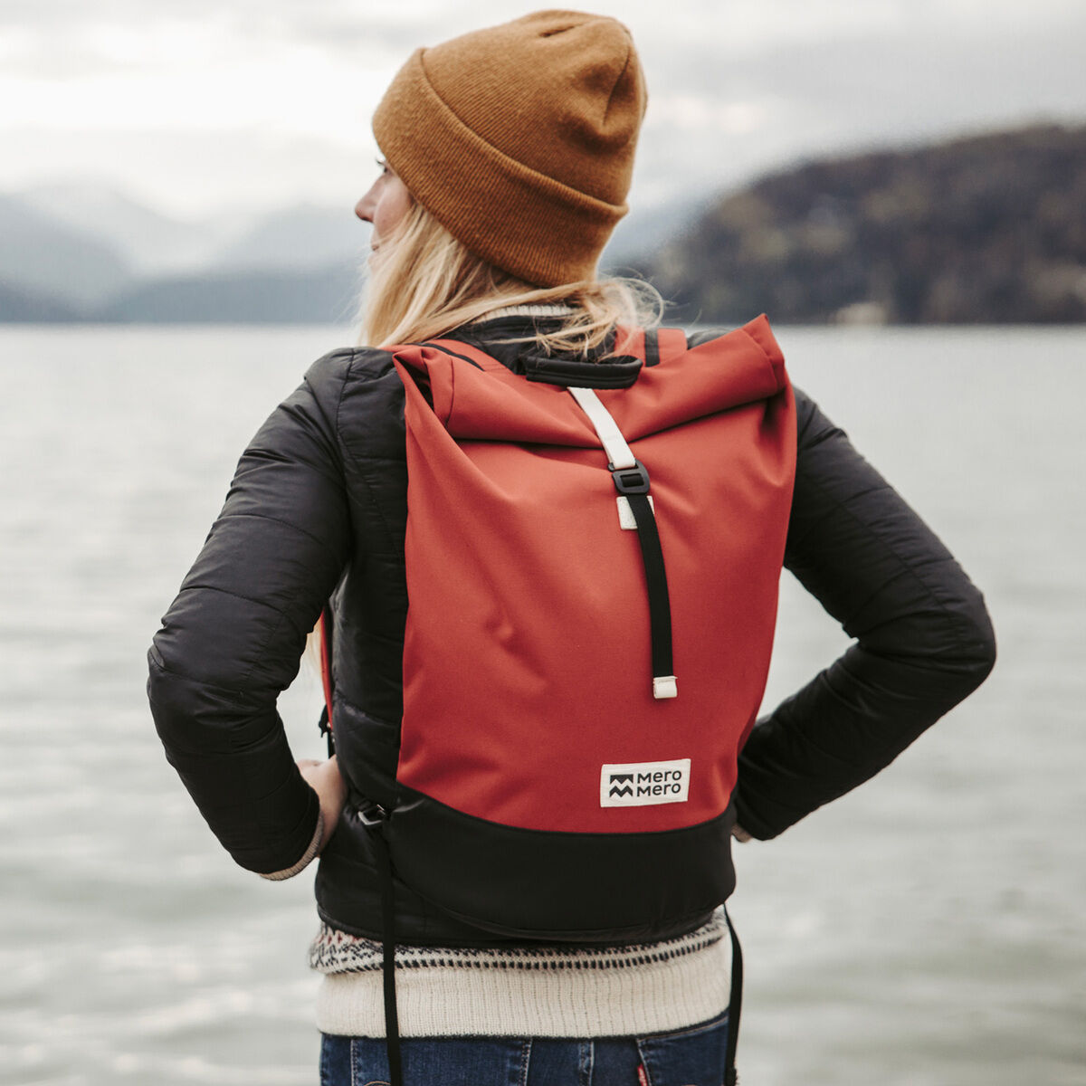Eine Frau steht vor einem See und trägt einen roten Rucksack »Wanaka«. Die Landschaft im Hintergrund besteht aus Wasser und Bergen.