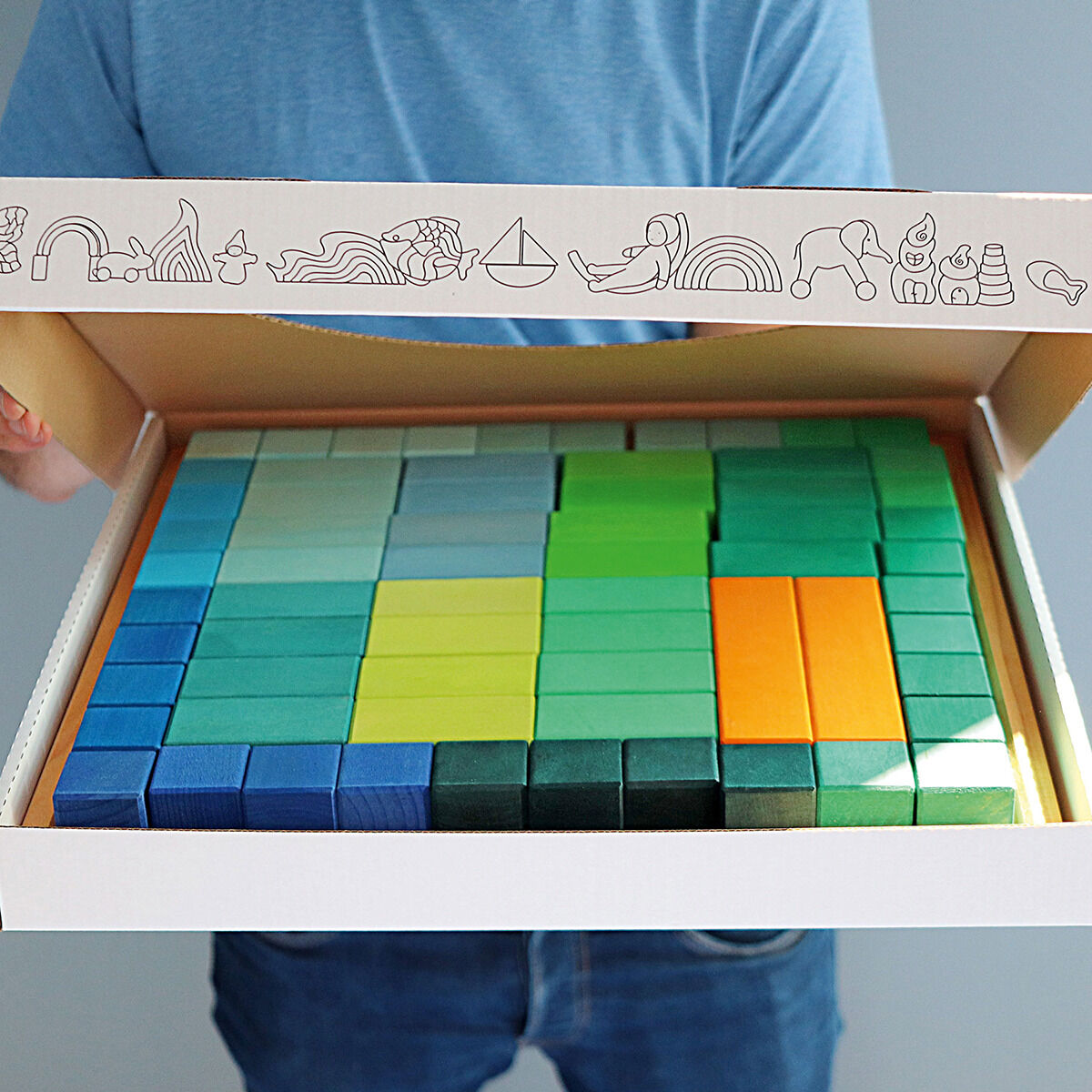 Ein buntes Set aus Holzbausteinen in Form einer Stufenpyramide, präsentiert in einer offenen Box. Das Spielzeug besteht aus verschiedenen quadratischen und rechteckigen Blöcken in Blau-, Grün-, Gelb- und Orangetönen. Diese kreativen Bausteine eignen sich besonders für Kinder und fördern das Lernen und die Motorik. Die Box ist mit dekorativen Symbolen verziert.