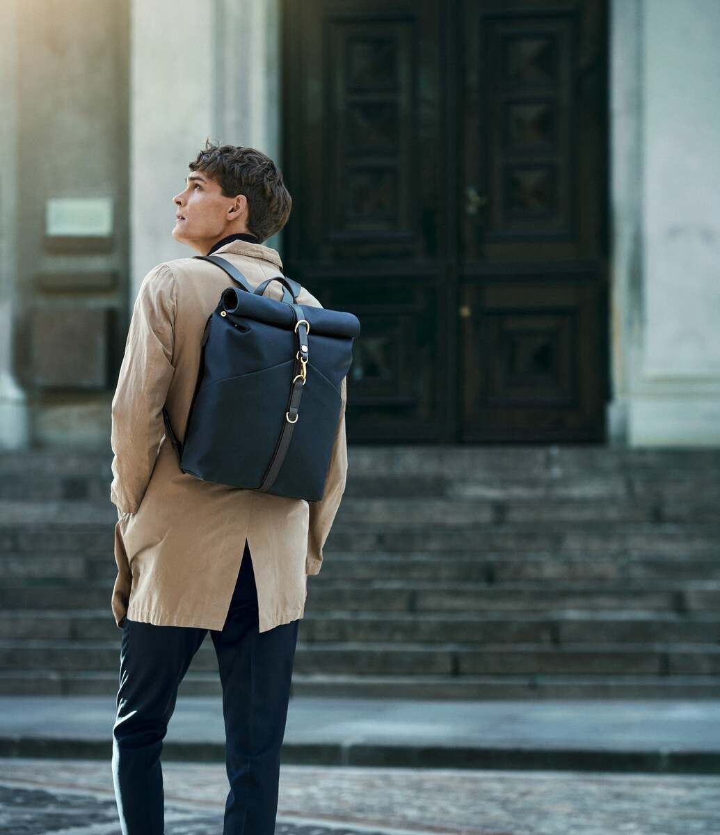 Ein Mann trägt einen eleganten, schwarzen Rucksack von MISMO und steht vor einem großen, alten Gebäude. Er trägt einen beigefarbenen Mantel und dunkle Hosen. Der Rucksack wirkt robust und stilvoll.
