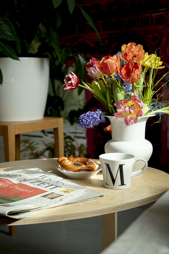 Auf einem runden Holztisch stehen eine weiße Frühstückstasse mit einem schwarzen Buchstaben 'M', eine Tageszeitung, ein Brezel und ein weißer Krug mit einem bunten Blumenstrauß. Im Hintergrund sind grüne Pflanzen sichtbar.