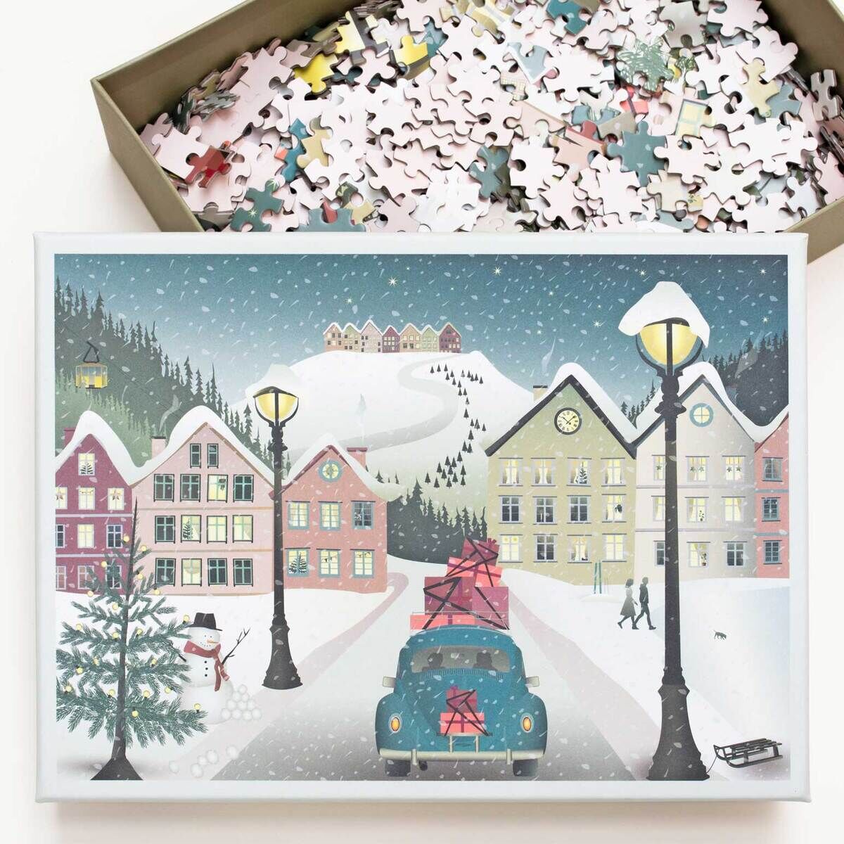Das Bild zeigt ein 1000-Teile Puzzle mit einer winterlichen Szene: Ein verschneites Bergdorf mit Häusern, dekorativen Straßenlaternen, einem Auto mit Geschenken auf dem Dach und spielenden Kindern. Der Hintergrund zeigt schneebedeckte Hügel und einen winterlich bewölkten Himmel. Das Puzzle liegt zum Teil in geöffneter Schachtel daneben.