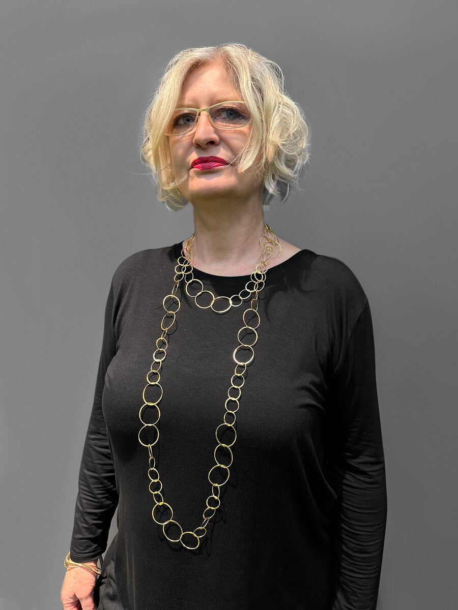 Eine Frau mit blonden, kurzen Haaren trägt eine lange Kette mit runden Elementen aus vergoldetem Silber. Sie trägt ein schwarzes Oberteil vor einem grauen Hintergrund.