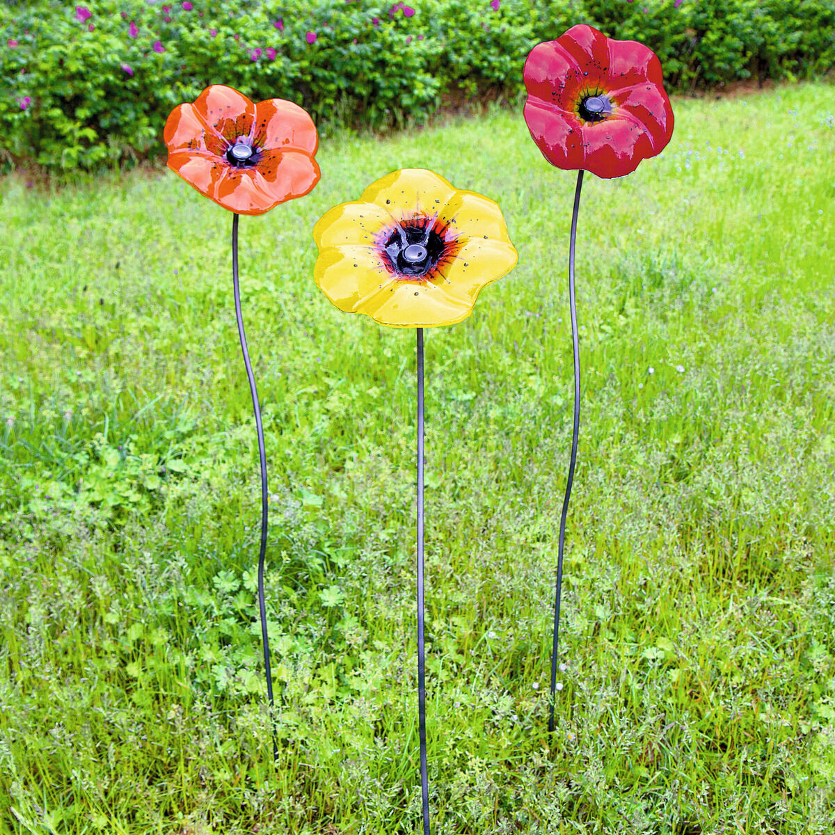 Drei farbenfrohe Gartenstecker aus Metall in Form von Blumen, in den Farben Rot, Gelb und Orange, stehen auf einer grünen Wiese vor einem Hintergrund mit Sträuchern. Geeignet als wetterfeste Dekoration für Garten oder Balkon.