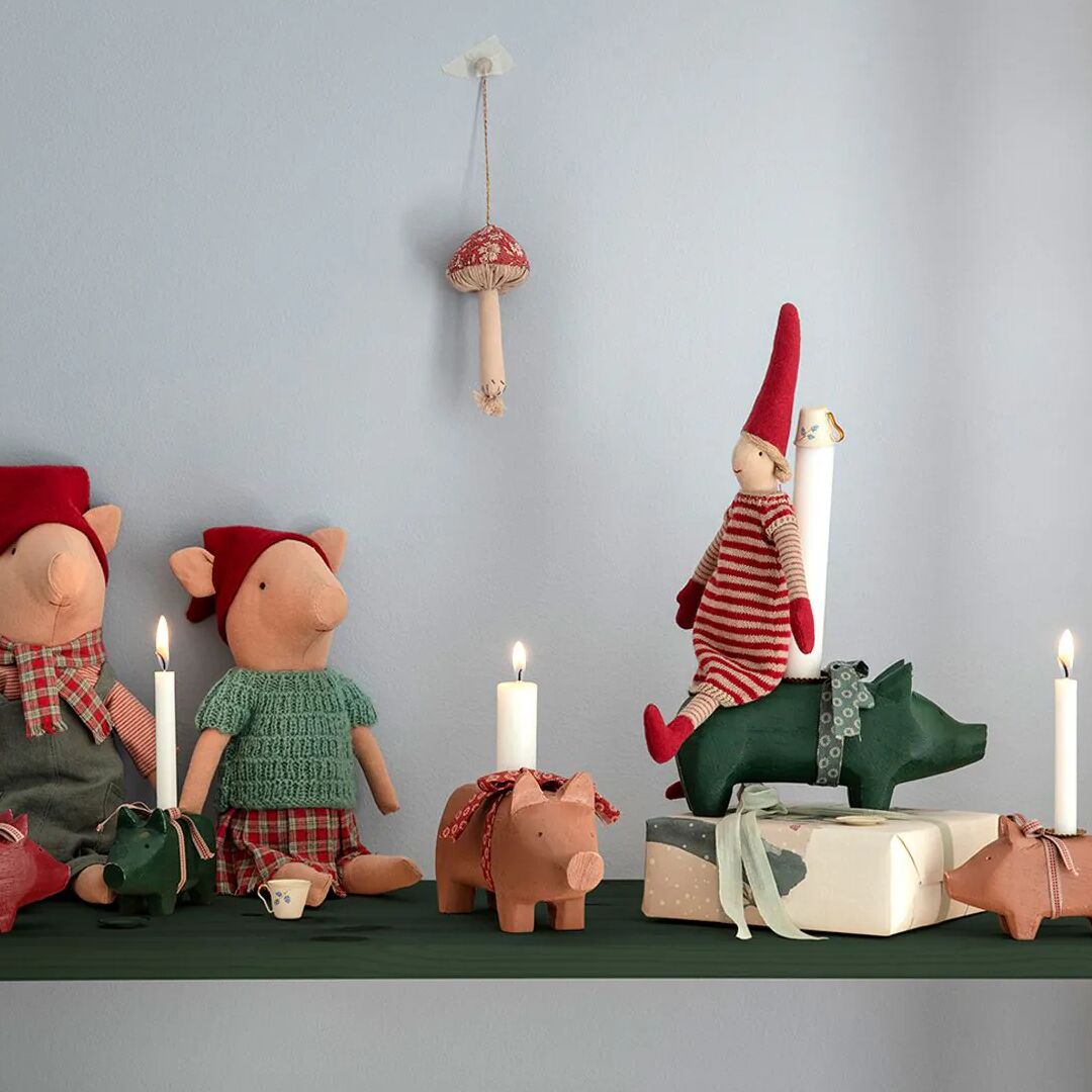 Mittlerer, grüner Kerzenhalter in Schweineform, platziert auf einem Regal neben weiteren Schweinefiguren, Kerzen und Geschenkboxen in gemütlicher, weihnachtlicher Dekoration. Im Hintergrund sitzen Stofffiguren in roter Kleidung vor einer hellblauen Wand.
