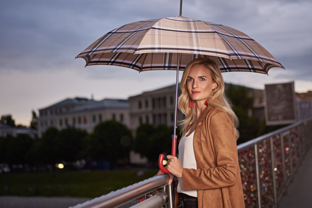 Blonde Frau im Freien, die bei bewölktem Himmel auf einer Brücke unter einem eleganten Schirm mit Ledergriff steht. Der Schirm hat ein beiges Plaidmuster.