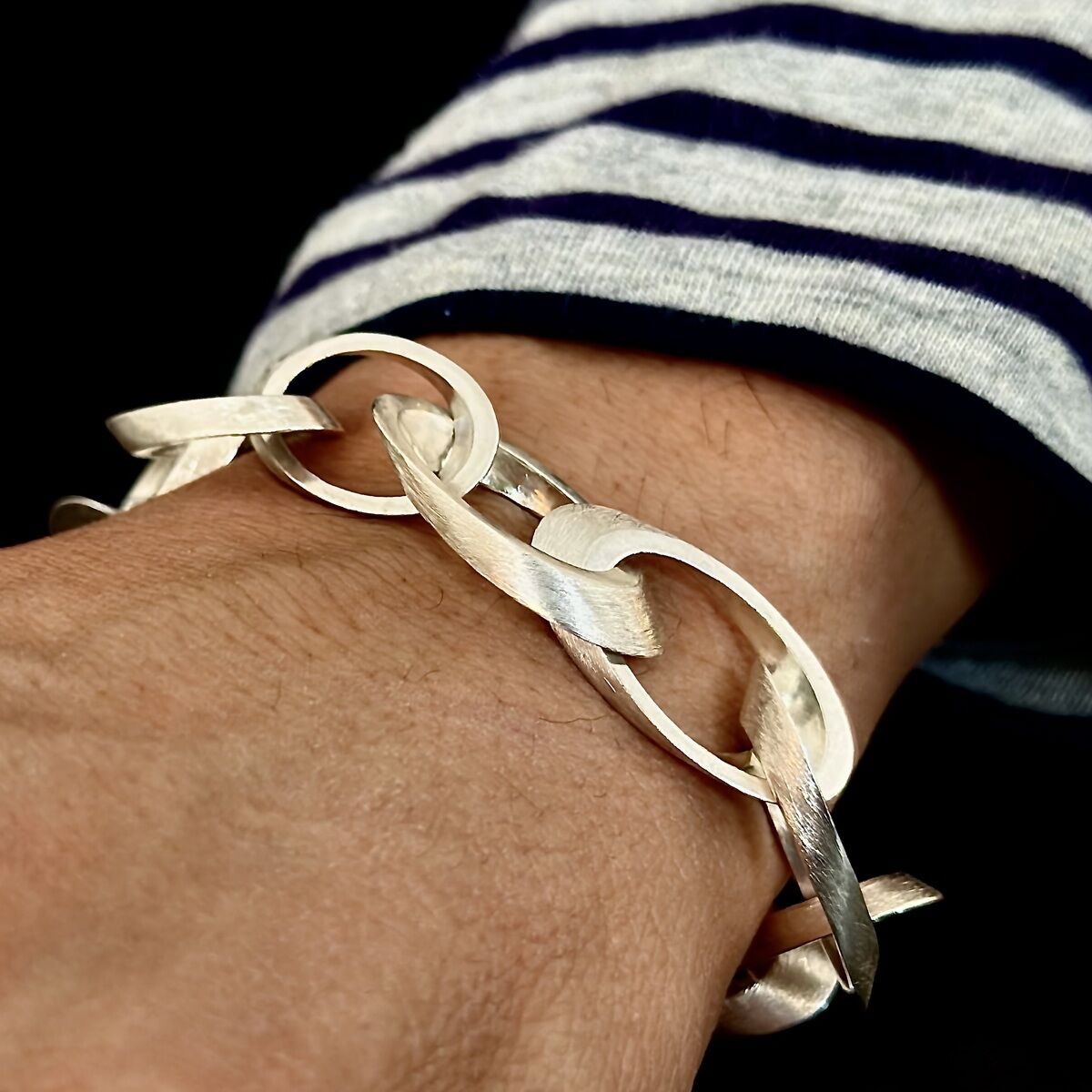 Ein silbernes Armband mit großen, ineinandergreifenden Kettengliedern an einem Handgelenk, getragen über einem gestreiften, dunkelblauen und weißen Stoff.