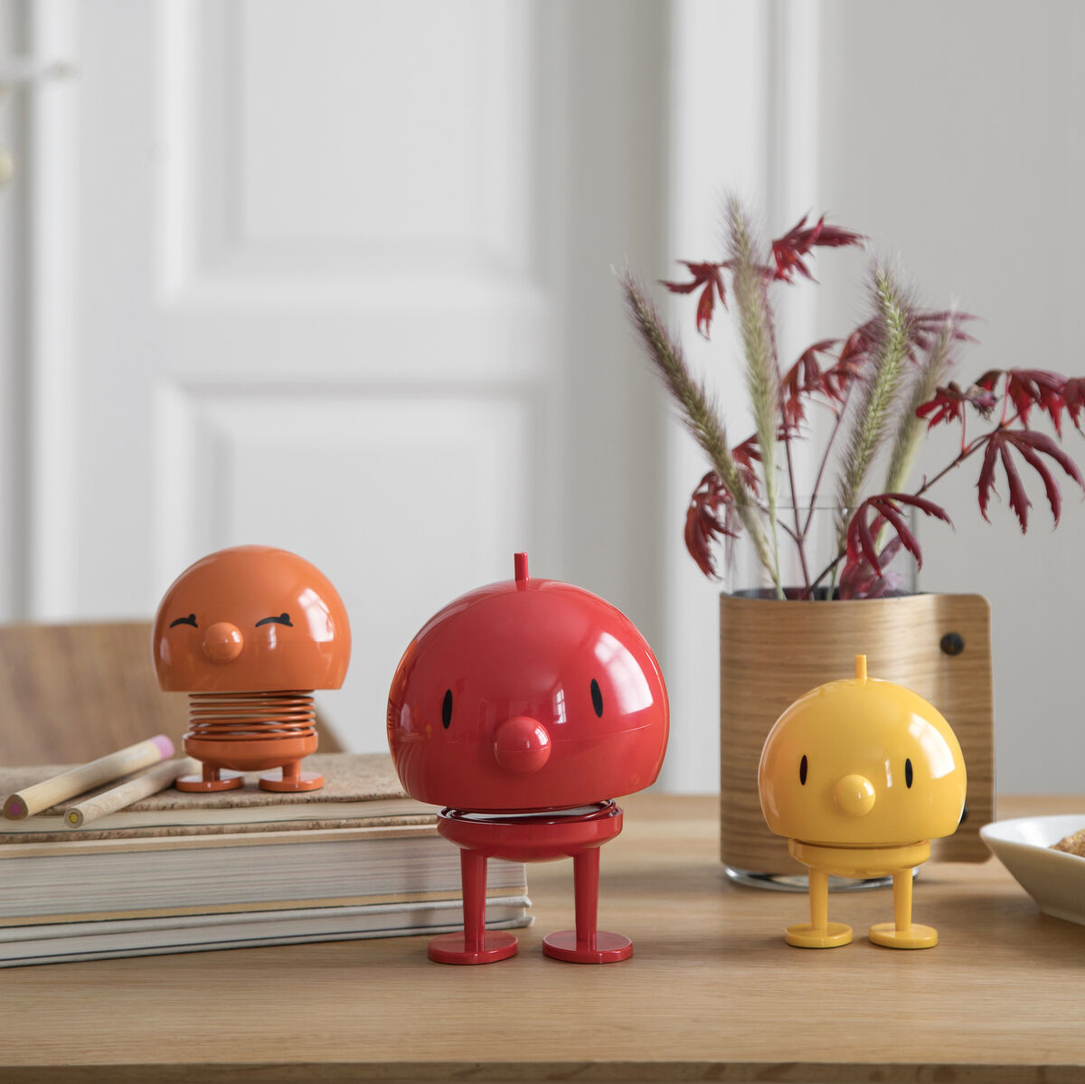 Drei fröhliche Hoptimist-Bumble Figuren in den Farben Rot, Orange und Gelb stehen auf einem Holztisch. Im Hintergrund sind Bücher, eine Pflanze in einer Vase und eine Schale mit Gebäck zu sehen.