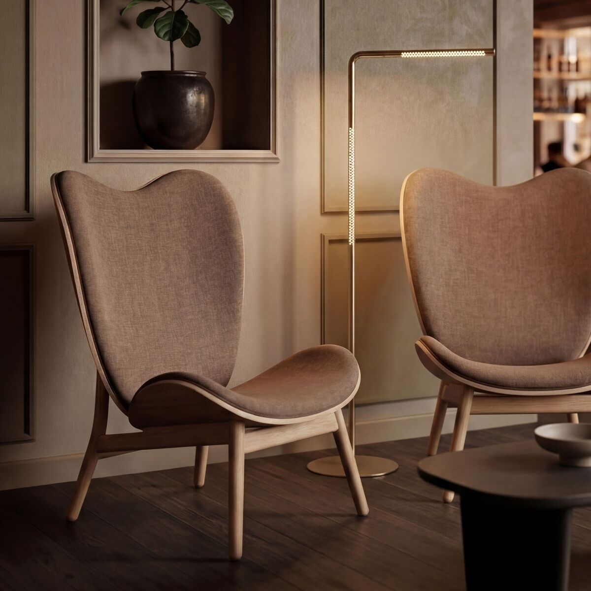 Stilvolles Wohnzimmer mit zwei Designerstühlen in Beige, einem schwarzen Tisch und der Stehleuchte »Omni« von Umage aus Messing. An der Wand steht eine Topfpflanze in einer Nische, der Raum ist in warmem Licht und Naturtönen gehalten, moderner skandinavischer Stil.