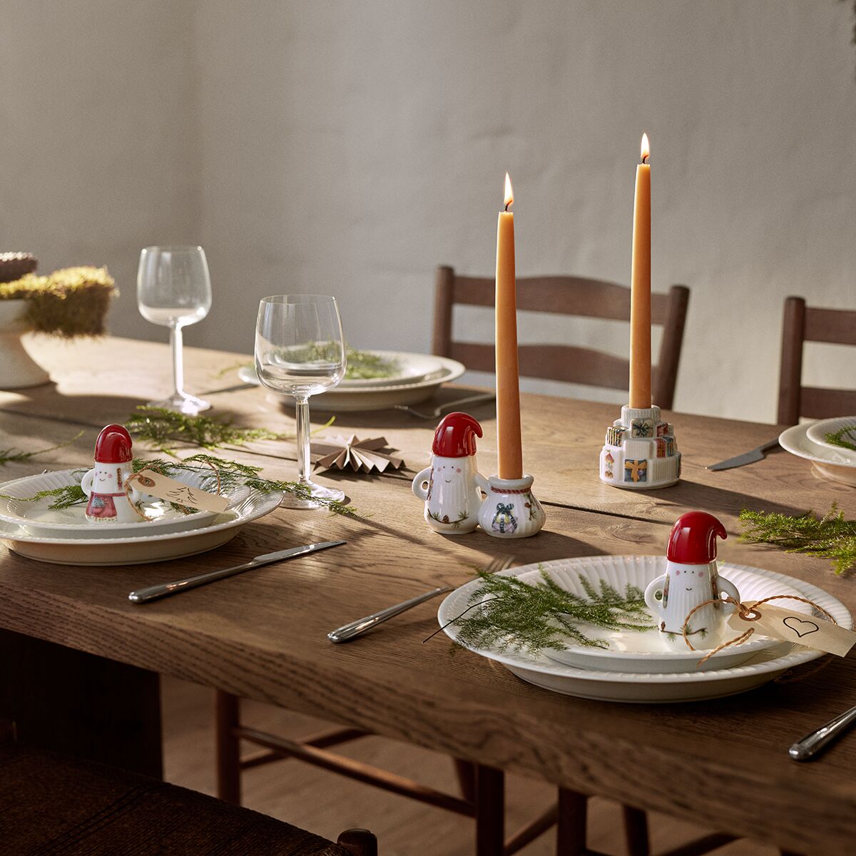 Ein festlich gedeckter Holztisch mit Keramikfiguren des Weihnachtsmanns aus der Hammershøi Kollektion, weißen Tellern mit grünem Tannenzweig, brennenden Stabkerzen und edlem Glasgeschirr. Die Szene vermittelt eine weihnachtliche und gemütliche Atmosphäre, ideal zur Advents- oder Weihnachtszeit.