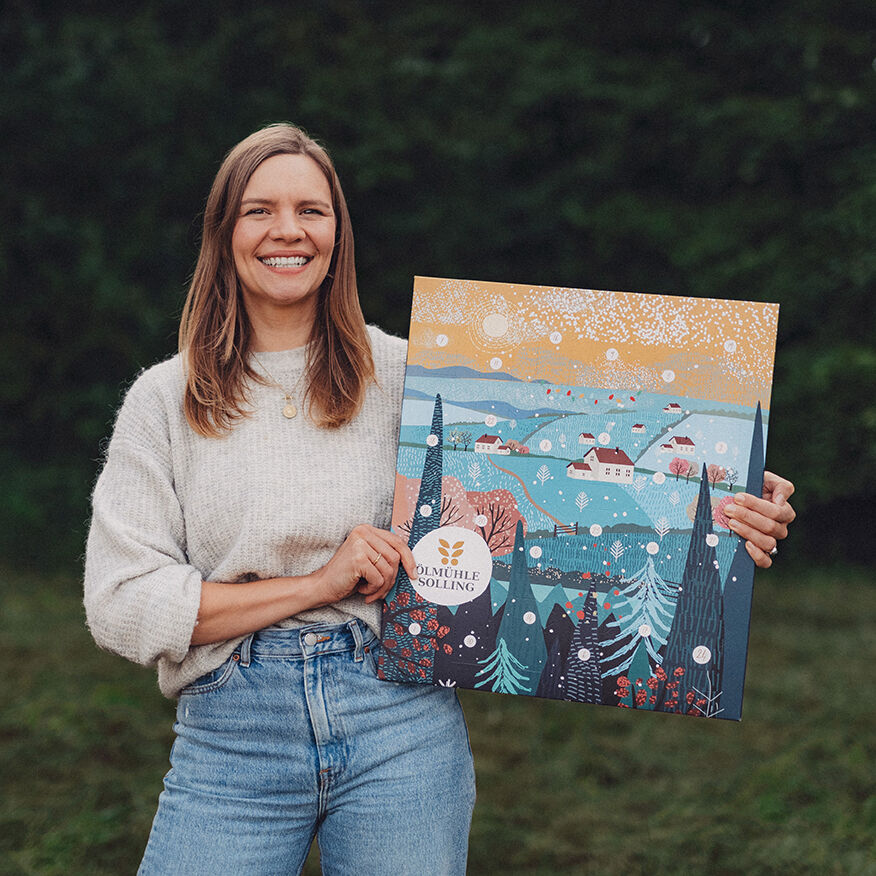 Frau im Freien hält den Adventskalender »Winterlandschaft« von Ölmühle Solling in den Händen. Der Kalender zeigt eine detailreich illustrierte, verschneite Winterlandschaft mit Häusern, Bäumen und Sternenhimmel auf kräftig buntem Design.
