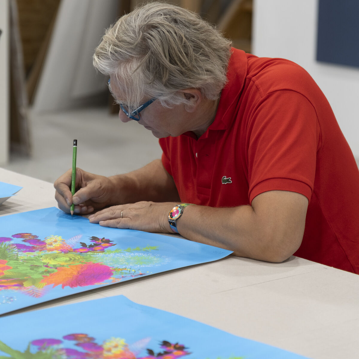 Miguel Chevalier arbeitet an einem blauen Kunstwerk mit bunten Naturmotiven auf einem Tisch. Der Künstler trägt ein rotes Polohemd und sitzt in einem Atelier.