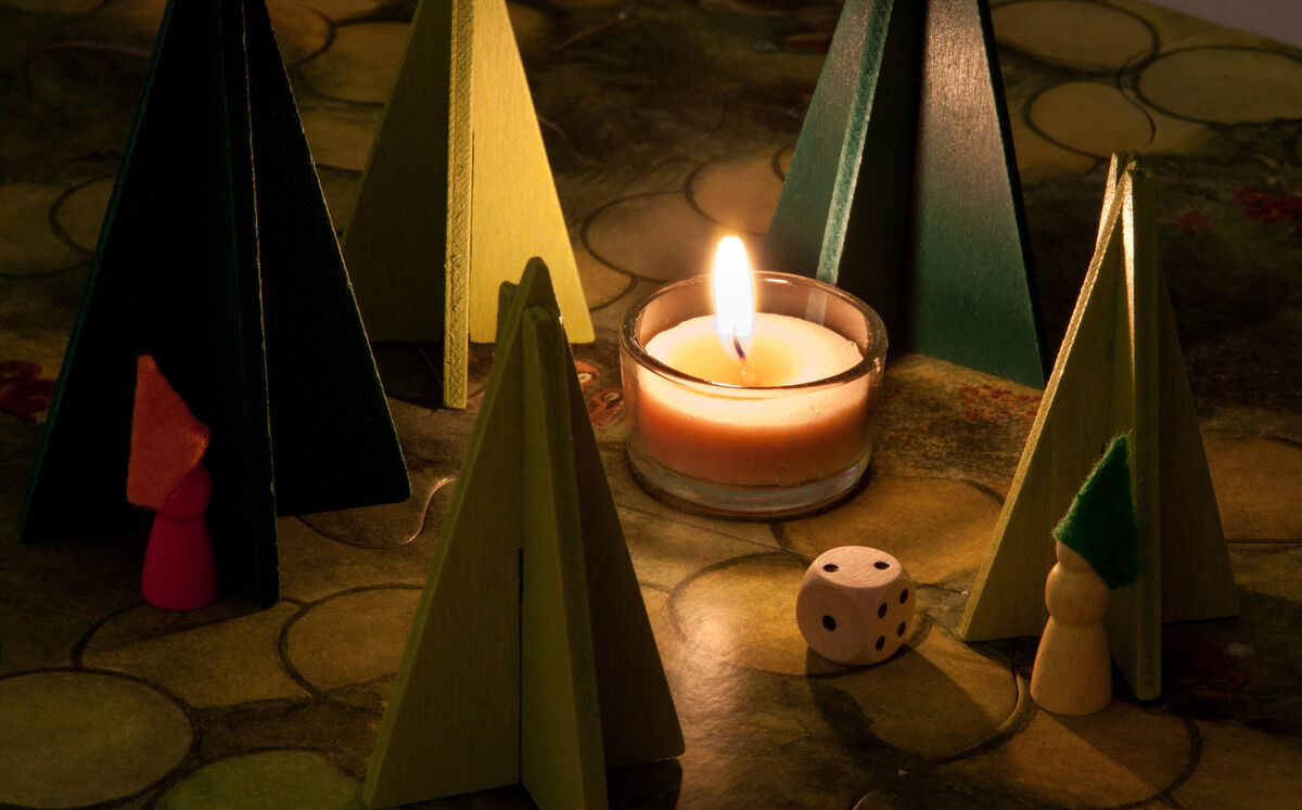 Das Bild zeigt das Brettspiel 'Waldschattenspiel', das auf einem Tisch aufgebaut ist. Verschiedene hölzerne Tannenbaum-Figuren stehen um eine brennende Kerze herum. Ein Würfel liegt ebenfalls auf dem Spielfeld und einige bunte Spielfiguren sind sichtbar.
