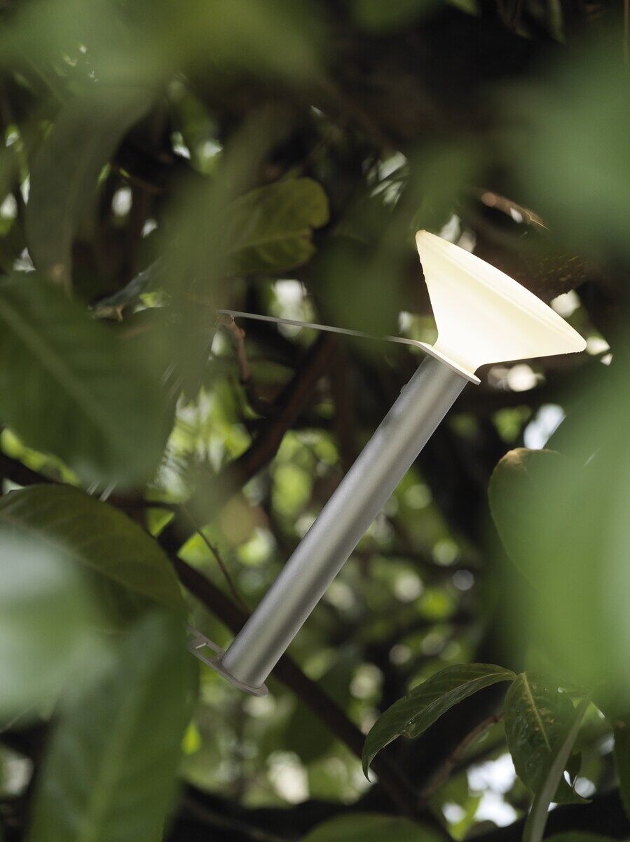 Eine tragbare Lampe »Piton« schwebt zwischen dichten grünen Blättern. Die Lampe hat einen silbernen Körper und strahlt ein warmweißes Licht aus. Ideal für Outdoor-Abenteuer wie Camping oder Gartenpartys.