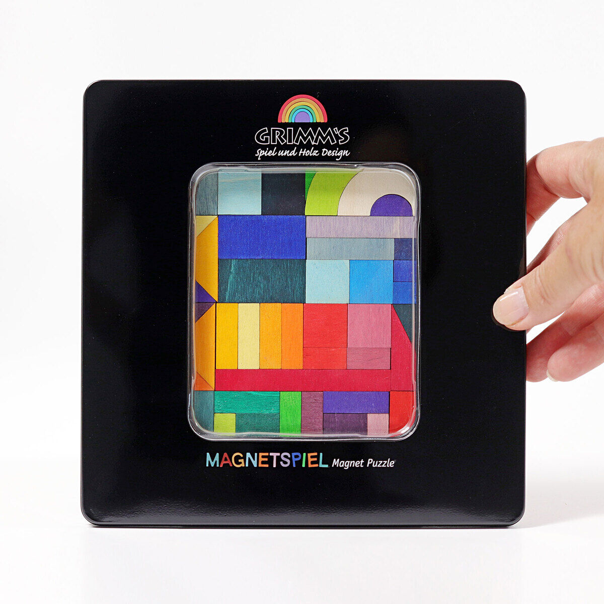 Magnetspiel »Geo-Grafisch« von Grimm's, ein buntes Holzpuzzle mit geometrischen Formen in einer schwarzen Verpackung, die von einer Hand gehalten wird.