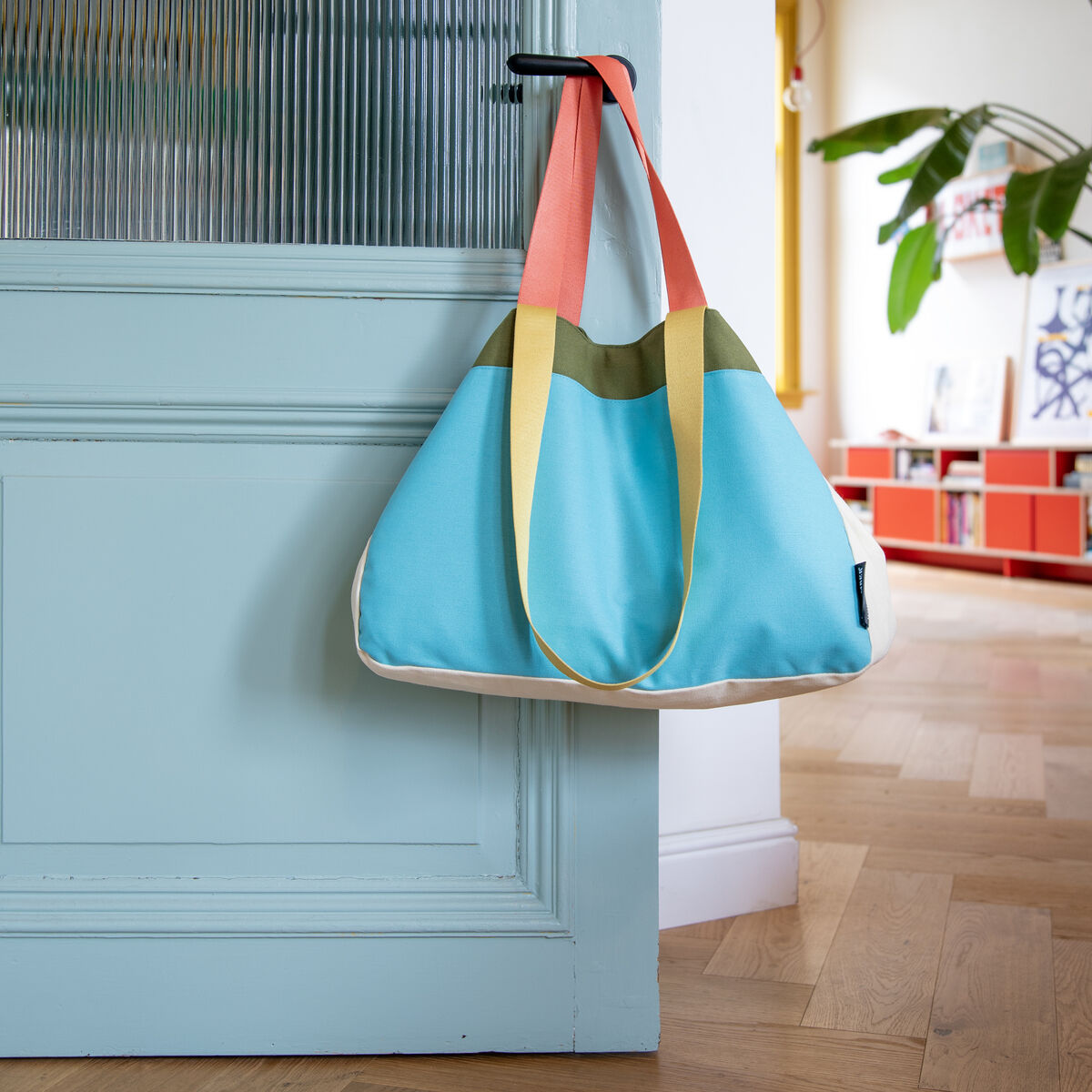 Bunte Canvas Tasche mit blauem Hauptteil, gelben und orangefarbenen Henkeln, vor hellblauer Tür in einer modernen Wohnung.
