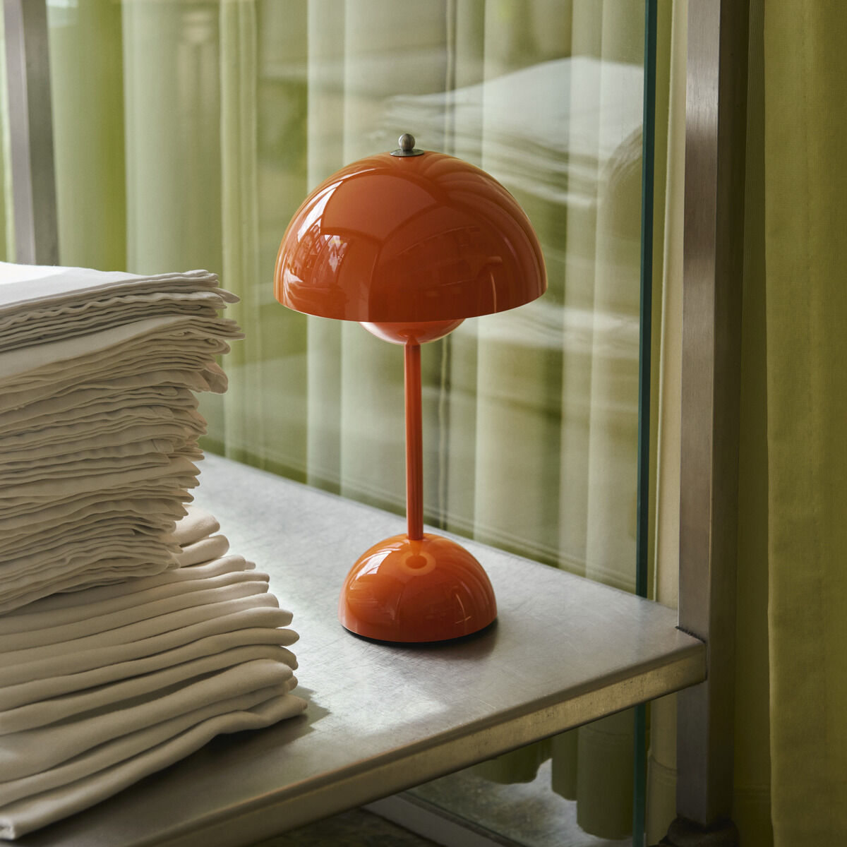 Auf einem metallischen Regal neben einem Stapel weißer Textilien steht eine orangefarbene Flowerpot VP9 Akku-Tischleuchte von &Tradition. Im Hintergrund hängen grüne Vorhänge, die den modernen und stilvollen Wohnraum ergänzen.