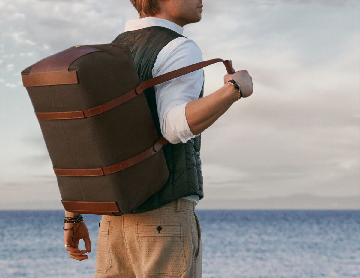 Ein Mann trägt die Reisetasche »SUPPLY« über der Schulter. Die Tasche besteht aus braunem Material mit Lederdetails. Im Hintergrund ist das Meer und ein heller Himmel zu sehen.