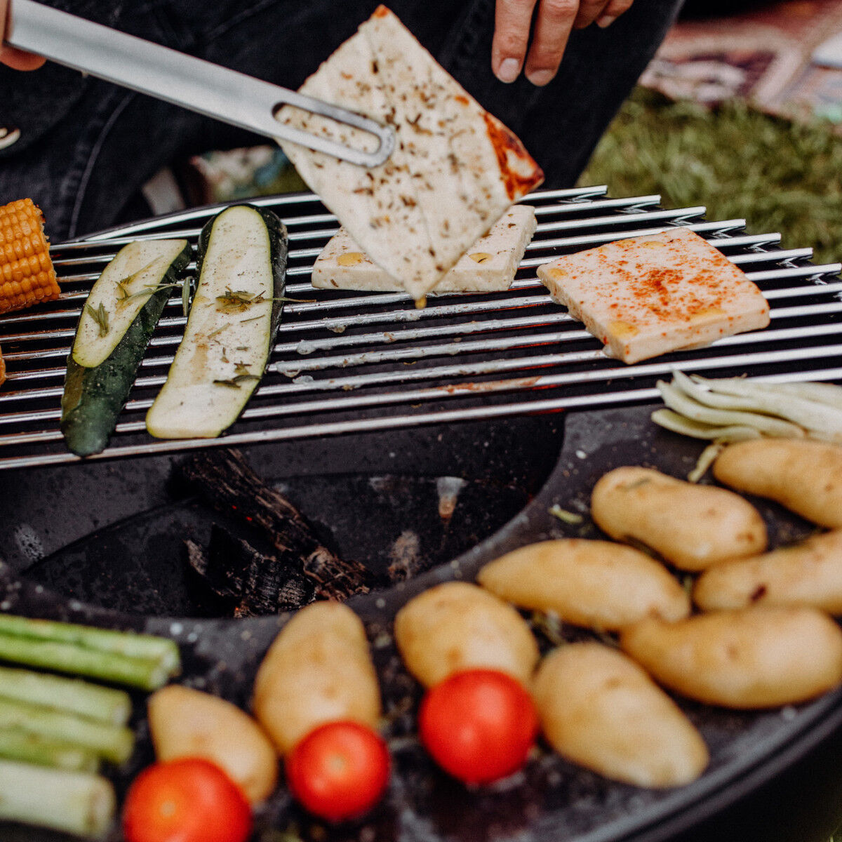 Eine Nahaufnahme eines Grills mit verschiedenen Gemüsearten darauf, wie Maiskolben, Zucchini, Tomaten und Kartoffeln. Eine Hand hält ein Grillspatel, der ein Stück Käse wendet. Es ist grüne Wiese und ein Picknick-Teppich im Hintergrund zu sehen.