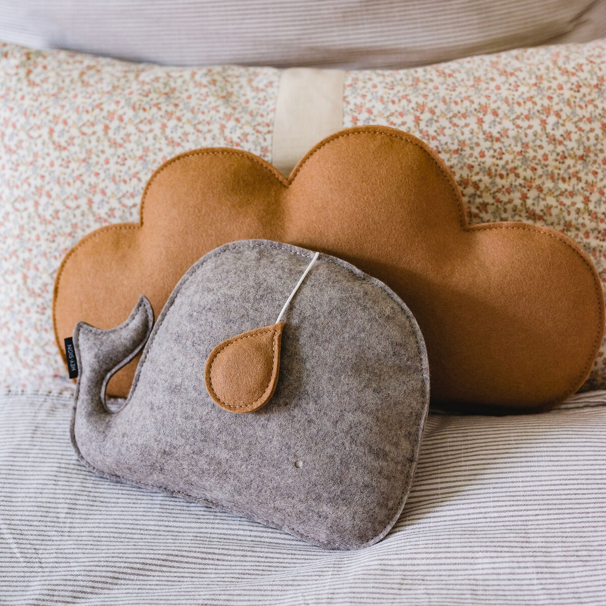 Eine graue Spieluhr in Form eines Wals liegt auf einem gestreiften Stoff, kombiniert mit einem braunen Kissen in Wolkenform im Hintergrund.