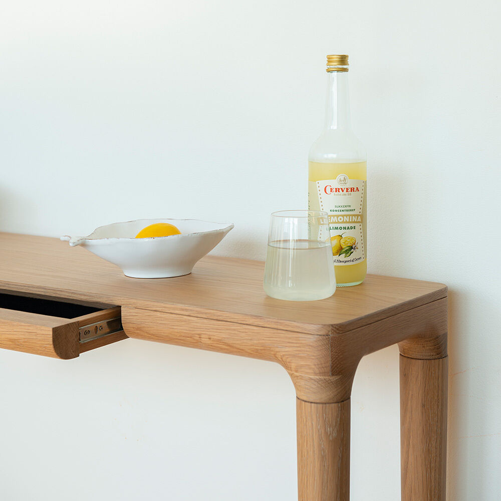 Heller Konsolentisch »Heart'n'Soul« aus massiver Eiche von Umage, im skandinavischen Stil mit abgerundeten Kanten. Auf dem Tisch stehen eine halbvolle Flasche Limonade, ein Glas und eine Schale mit einer halben Zitrone, daneben eine leicht geöffnete Schublade.