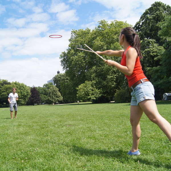 Zwei Personen spielen auf einer großen, grünen Wiese »Tualoop«. Eine Frau wirft mit Stäben einen Ring, während ein Mann ihr gegenüber steht, um ihn zu fangen. Es ist ein sonniger Tag mit blauem Himmel und es gibt Bäume im Hintergrund.