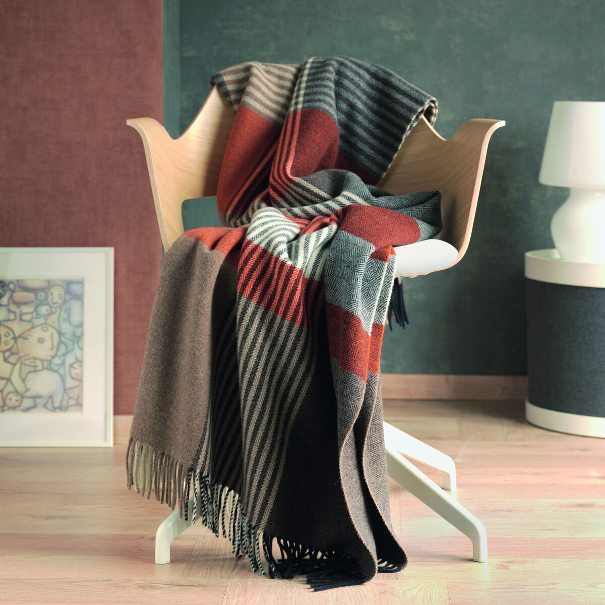 Eine dekorativ auf einem modernen Holzstuhl drapierte Plaiddecke namens »Harris« mit einem Streifenmuster in Braun-, Grau-, Rot-, Schwarz- und Weißtönen. Im Hintergrund sind eine Wand in Grau und Rosa sowie ein minimalistisches Kunstwerk und eine Lampe auf einem Beistelltisch zu sehen.