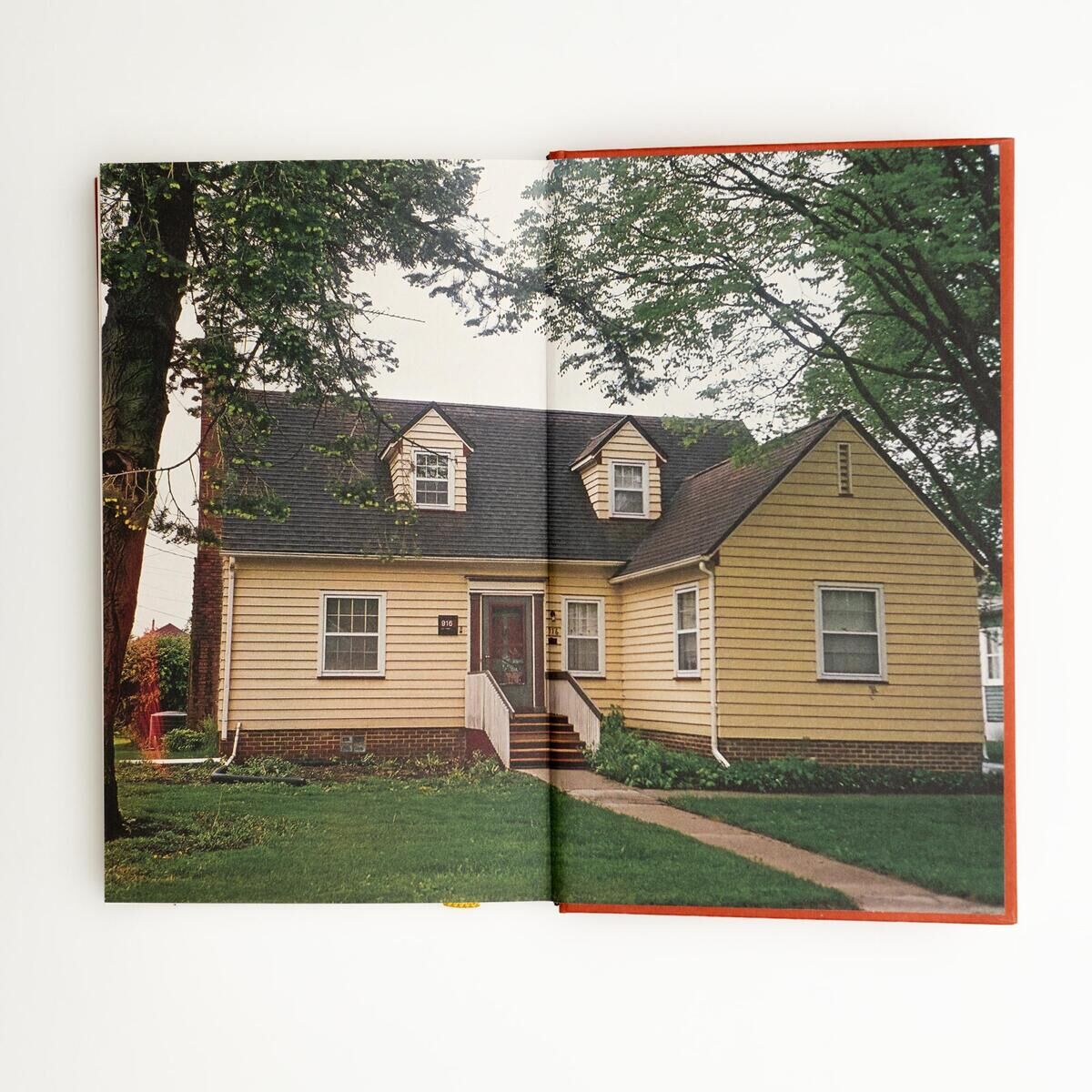 Offenes Buch mit einem Foto eines gelben Wohnhauses mit Satteldach, umgeben von grünem Rasen und großen Bäumen. Die Architektur und der gepflegte Vorgarten sind gut sichtbar. Das Bild hat einen künstlerischen und dokumentarischen Charakter.