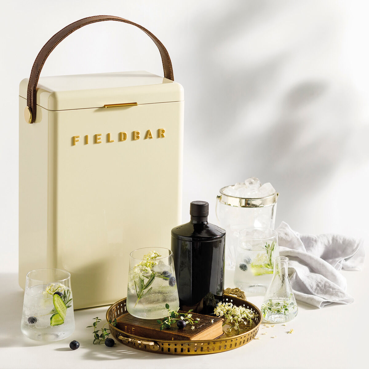 Eine cremeweiße Getränkebox der Marke Fieldbar mit braunem Griff steht auf einem weißen Hintergrund. Daneben befinden sich ein schwarzer Ginbehälter, Gläser mit klaren Getränken, Eiswürfel und Kräutergarnituren. Ein Tablett aus Messing und einige frische Beeren runden das Bild ab.