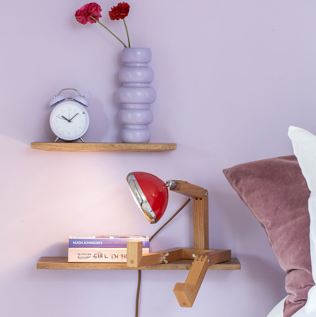 Ein modernes Schlafzimmer mit einer dekorativen Leuchte »Mr. Wattson« aus Holz und rotem Lampenschirm auf einem Regal. Daneben befinden sich ein Wecker, eine rosa Vase mit Blumen und Bücher. Die Wand und Bettkissen sind in Fliedertönen gehalten.