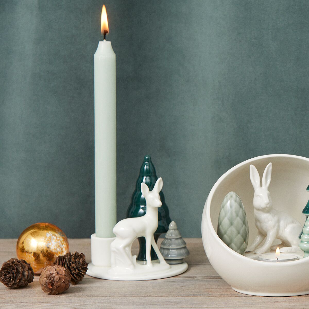 Auf einem Holztisch steht ein handgefertigtes Porzellan-Dekorationsset von Dottir Nordic Design: Ein weißes stehendes Bambi mit grünen, mattierten Tannenbäumen, das gleichzeitig als Kerzenhalter für eine helle Stabkerze dient. Daneben Tannenzapfen, eine goldene Weihnachtskugel und weiteres Porzellanobjekt mit Kaninchen und Tanne.
