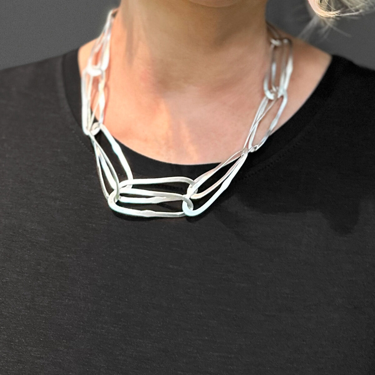 Eine elegante Frau mit blondem Haar trägt eine stilvolle Silberkette mit einem wellenförmigen Design. Der Hintergrund ist grau und die Frau trägt ein schwarzes Oberteil.