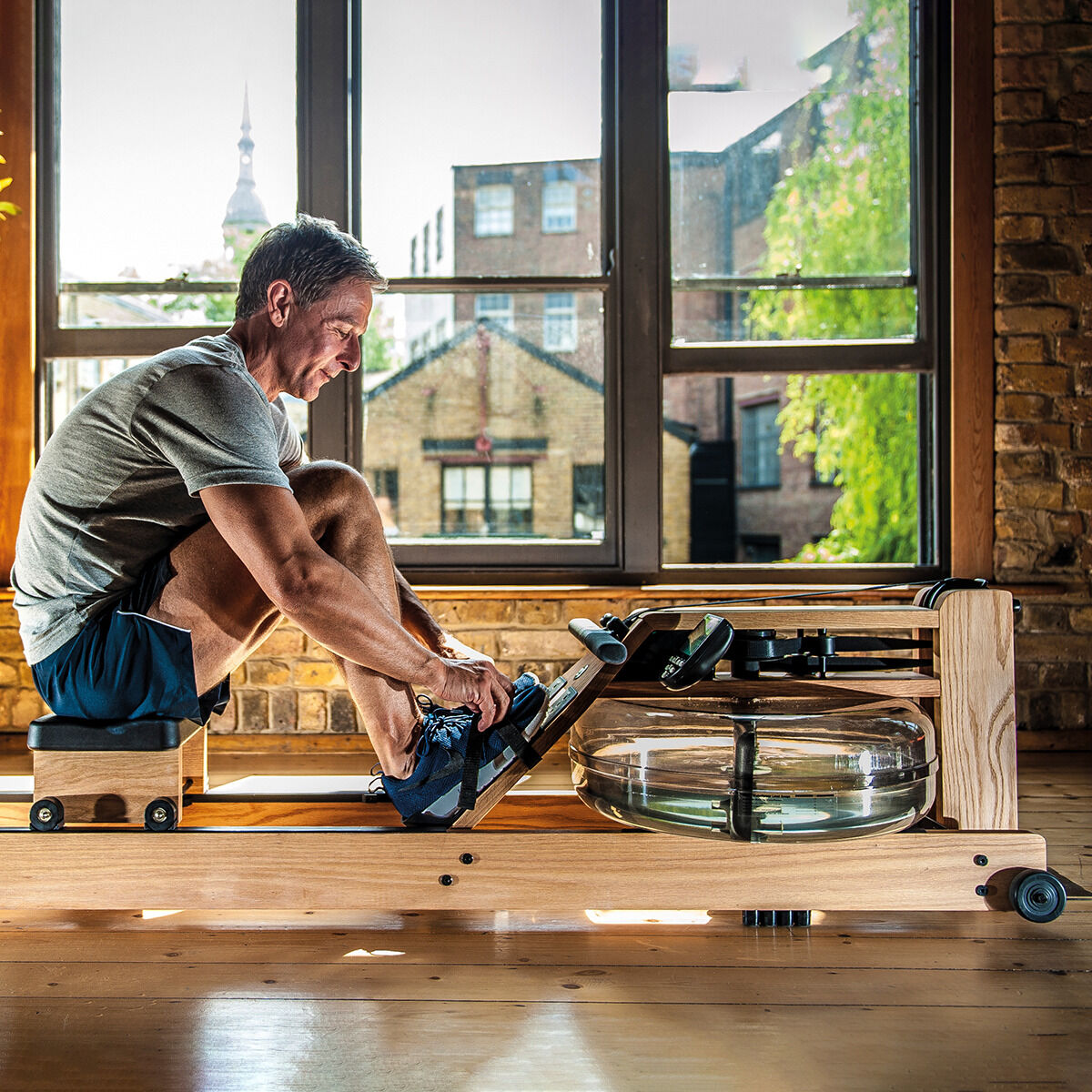 Mann trainiert auf einem Waterrower-Rudergerät aus Eiche, das in einem stilvollen Raum mit großen Fenstern steht. Das Gerät hat einen Holzrahmen und einen Wasserwiderstandstank. Es ist für Indoor-Cardio-Training geeignet.