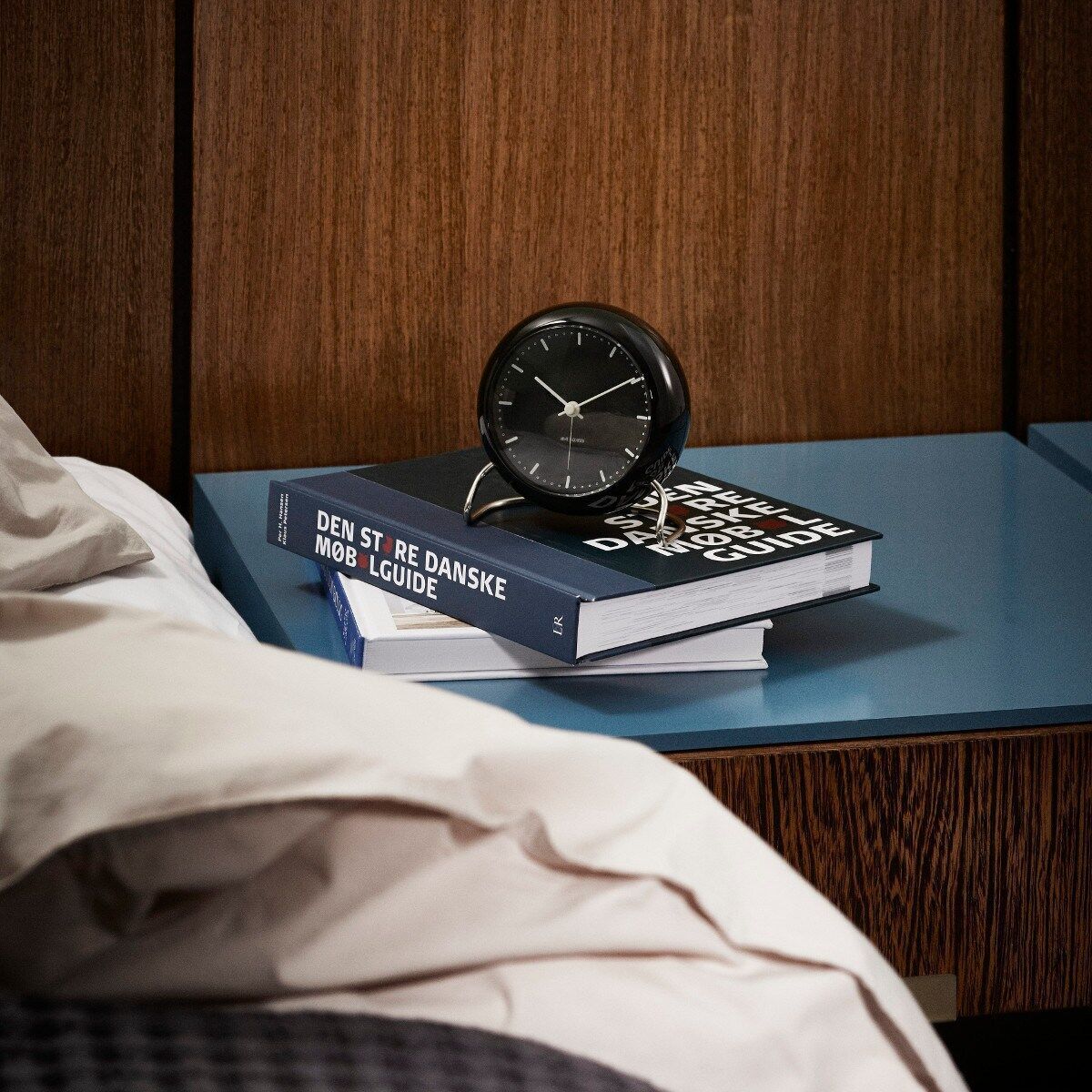 Eine stilvolle Tischuhr »AJ City Hall« in Schwarz von Arne Jacobsen, die auf einem blauen Tisch neben einem Buch steht. Die Szene befindet sich in einem gut eingerichteten Raum, wodurch das moderne und minimalistische Design der Uhr hervorgehoben wird.