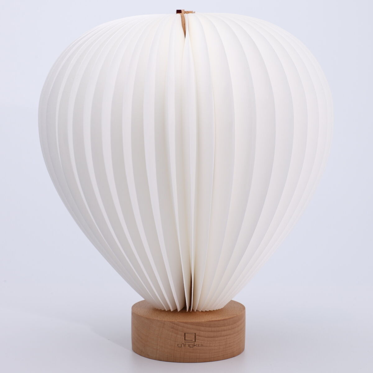 Auf dem Bild ist die formschöne Leuchte »Aeris Light« der Marke Gingko Design Ltd zu sehen. Die Lampe erinnert an einen stilisierten Heißluftballon, besteht aus weißen, lamellenartig gefalteten Elementen und steht auf einem runden Fuß aus hellem Eschenholz.