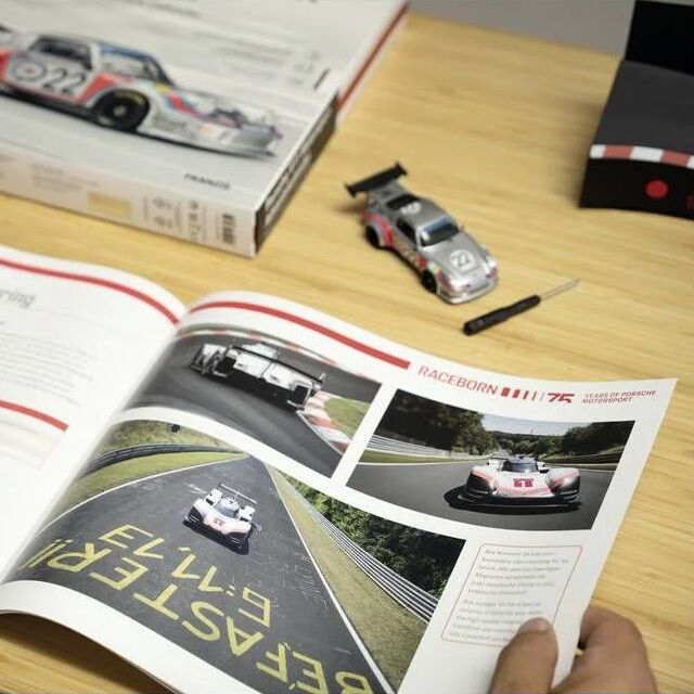 Auf einem Holztisch liegt ein geöffneter Bildband mit Motorsportfotos, darüber ein Modellauto Porsche 911 Turbo 1:43, ein kleiner Schraubendreher und die Verpackung vom Adventskalender »FRANZIS 67275 - Porsche 911 Turbo Motorsport Adventskalender 1:43«.