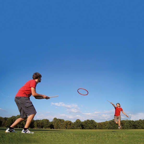 Zwei Personen spielen Tualoop auf einer grünen Wiese unter einem strahlend blauen Himmel. Beide sind in roter Kleidung und werfen einen Ring hin und her.