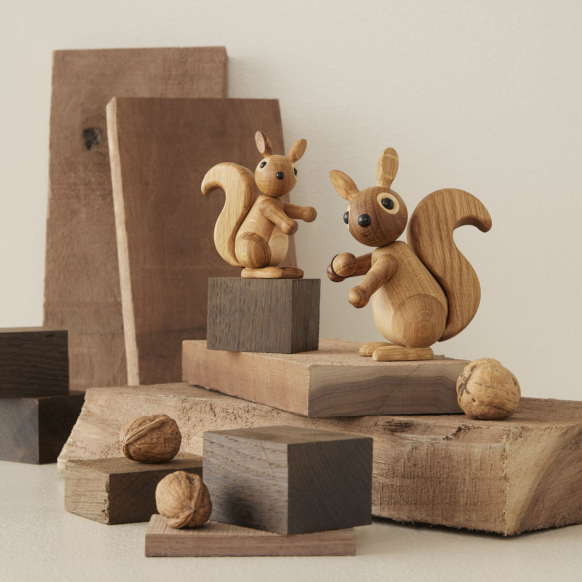 Zwei Eichhörnchen-Holzfiguren namens »Peanut« stehen auf gestapelten Holzstücken und Quadern, umgeben von Walnussschalen. Die Szene zeigt naturbelassene und verschiedenfarbige Holzoberflächen, die den natürlichen Look und handgefertigten Charakter hervorheben.