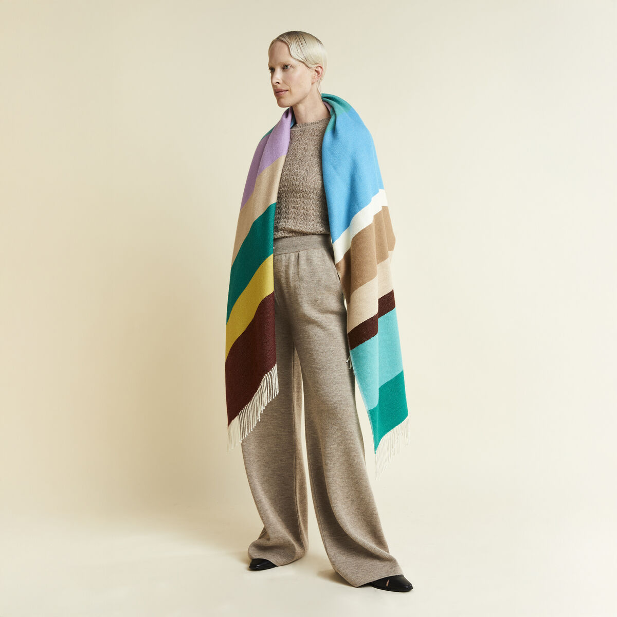 Eine Frau trägt das luxuriöse Alpaka-Cape »Mainau« von parati, das aus großflächigen Farbblöcken in Türkis, Grün, Gelb, Bordeaux, Pastelllila, Beige und Braun mit Fransen besteht. Das Cape wirkt weich, farbenfroh und modern zu eleganter Wollhose kombiniert.