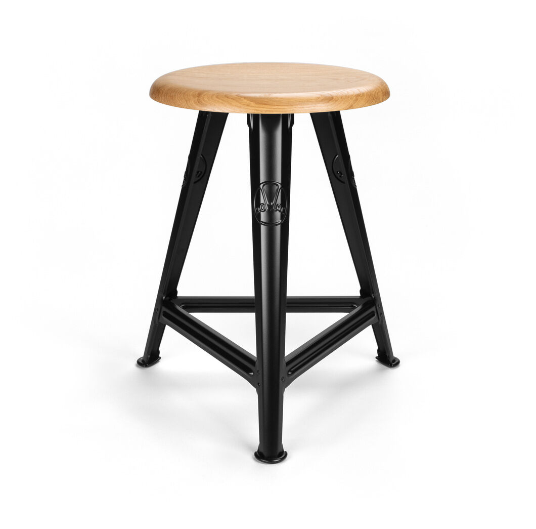 Ein industrieinspirierter Hocker mit einem runden Sitz aus Eichenholz und einem mattschwarzen Metallrahmen. Der Hocker steht stabil auf drei Beinen.